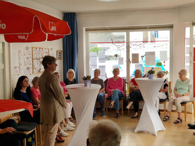 Frau Schmarsow steht an einem Tisch, rundum sitzen Gäste, in einem festlich geschmückten Raum.